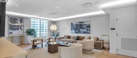Una sala de estar moderna con un juego de sofá, una mesa de centro y sillones. Amplias ventanas ofrecen luz natural y vistas de la ciudad.