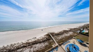 Blick auf einen Strand mit blauem Himmel, feinem Sand und einem Poolbereich mit Liegestühlen.