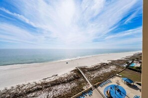 Blick auf einen Strand mit blauem Himmel, feinem Sand und einem Poolbereich mit Liegestühlen.