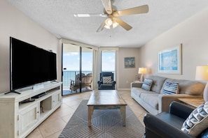 A living room with a television, a ceiling fan, a sofa, a coffee table, and a view of the ocean through a sliding glass door. - Phoenix 5 910 (Orange Beach)