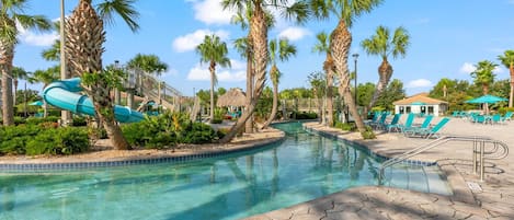 A pool area with clear water, palm trees, a water slide, lounge chairs, and a building in the background.