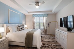 Une chambre à coucher comprenant un lit, des tables de chevet, un téléviseur, un ventilateur de plafond et une fenêtre avec des rideaux.