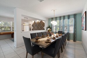 A dining area with a wooden table set for six, surrounded by dark chairs, with a decorative wall and a window with curtains. - Solara Resort 330 - Nine Bedroom Villa, Sleeps 22 (Orlando)