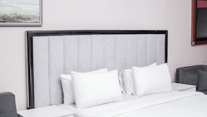 A hotel room with a bed, white bedding, a grey headboard, a nightstand, and a grey armchair. - JALITHS Boutique Hotel (Lusaka)