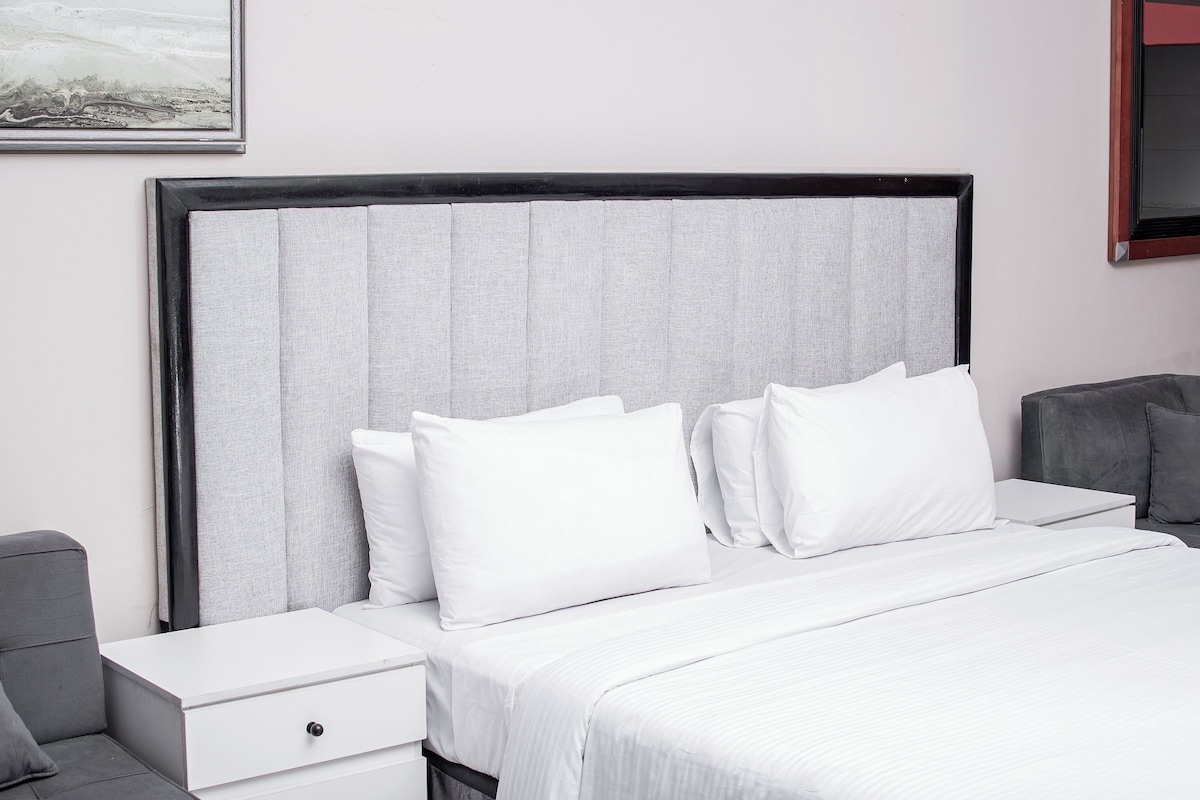 A hotel room with a bed, white bedding, a grey headboard, a nightstand, and a grey armchair.