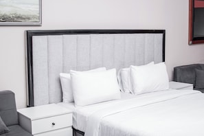 A hotel room with a bed, white bedding, a grey headboard, a nightstand, and a grey armchair. - JALITHS Boutique Hotel (Lusaka)