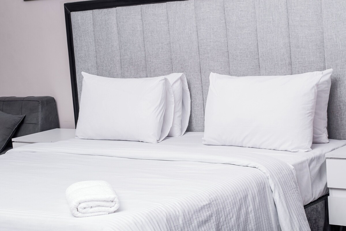 Habitación de hotel con una cama, dos almohadas, una toalla blanca doblada con cuidado y un cabecero gris.