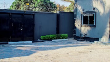 A gated entrance with a metal gate, a small green bush, and a building with a window and a door.