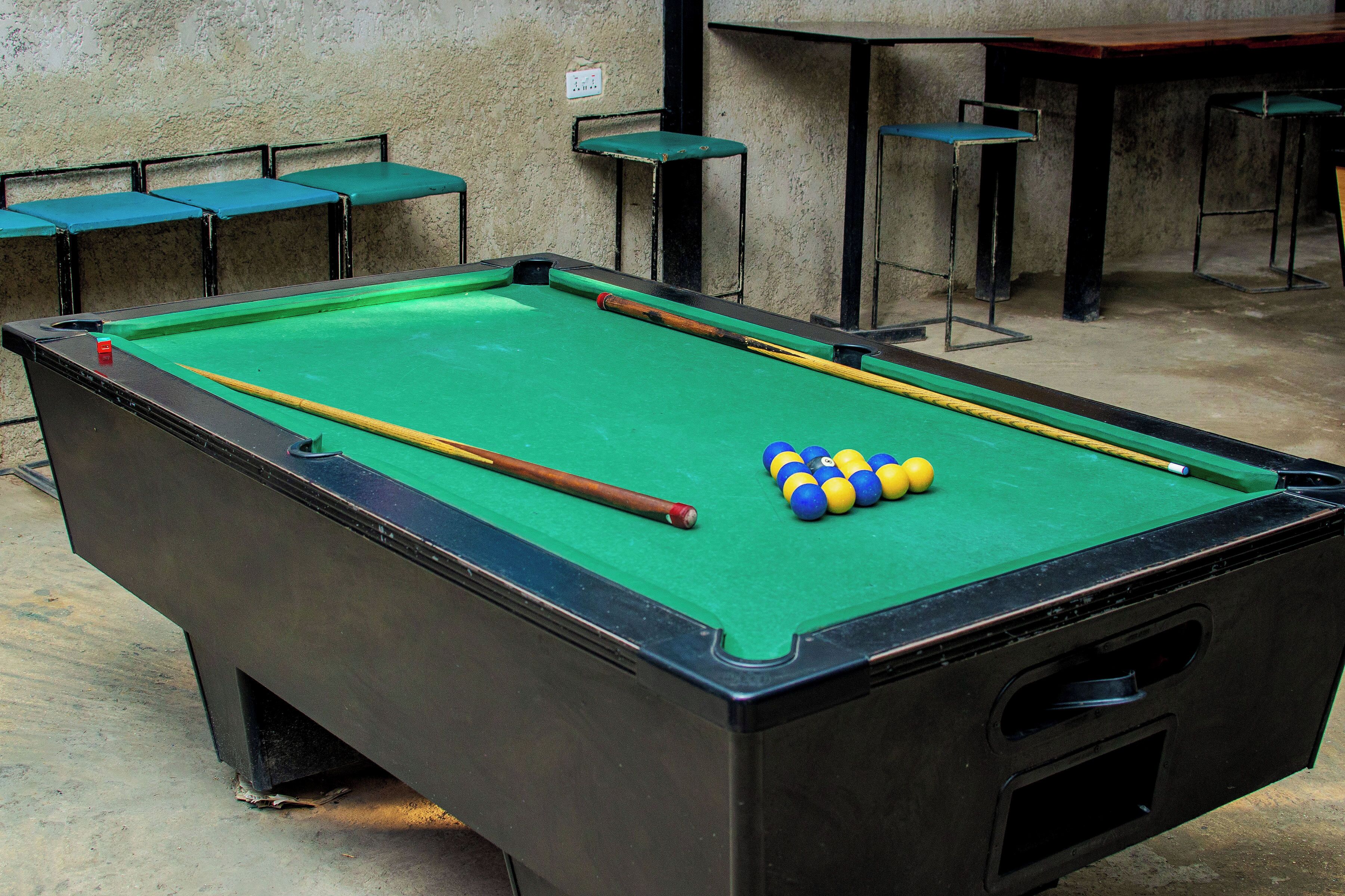 A pool table with a cue stick and balls, surrounded by green tables and chairs.