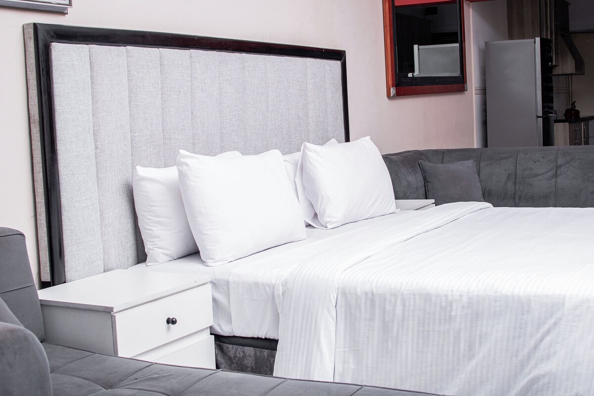 Una cama bien hecha con ropa blanca y almohadas, cabecera gris y mesita de noche blanca con un cajón.