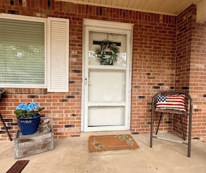 A brick house with a white door, a wreath, a blue flower pot, and a chair with an American flag pillow.