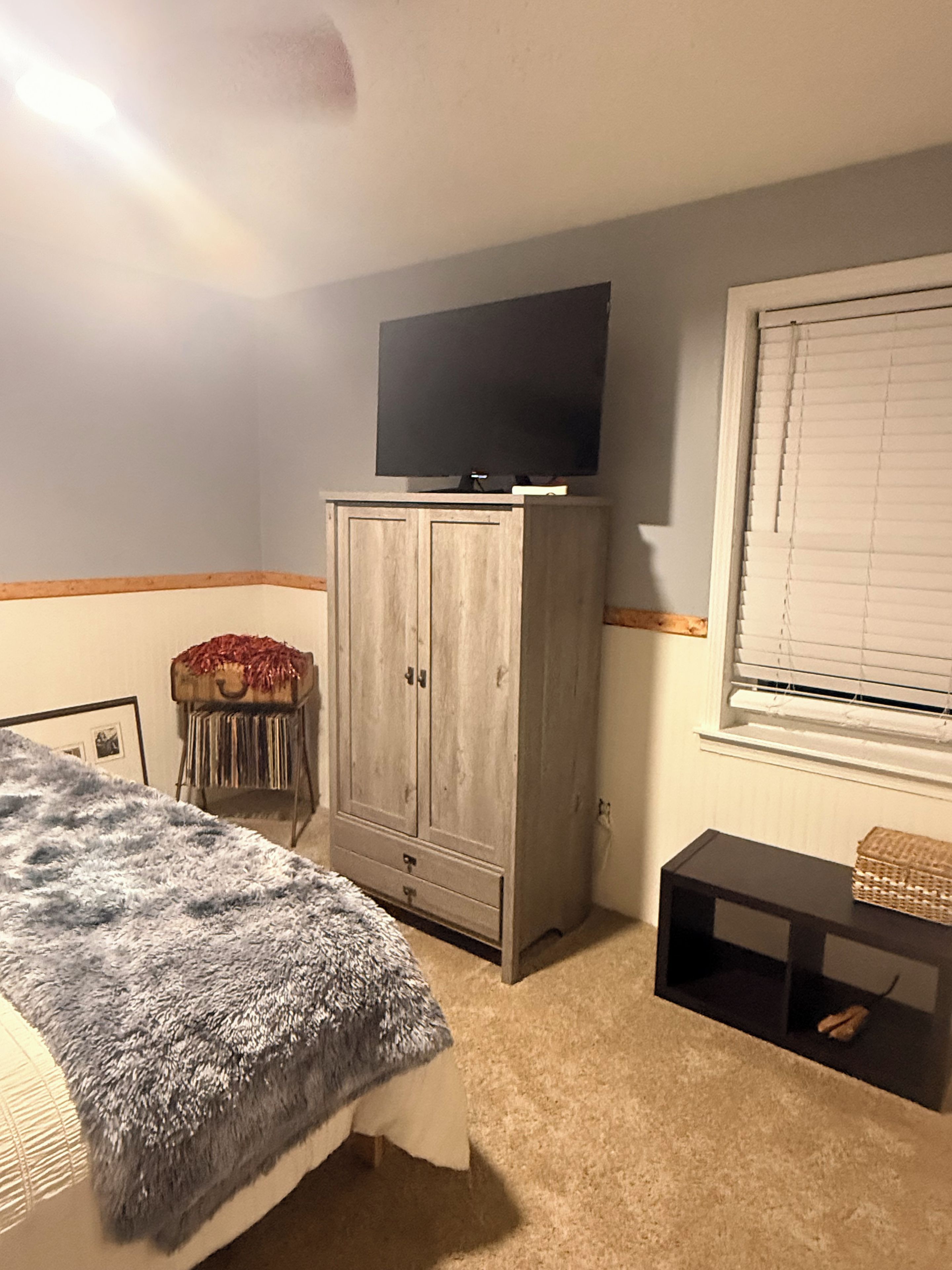 Un dormitorio con cama, armario, televisor encima, ventana con persianas y una mesita de noche con una cesta.