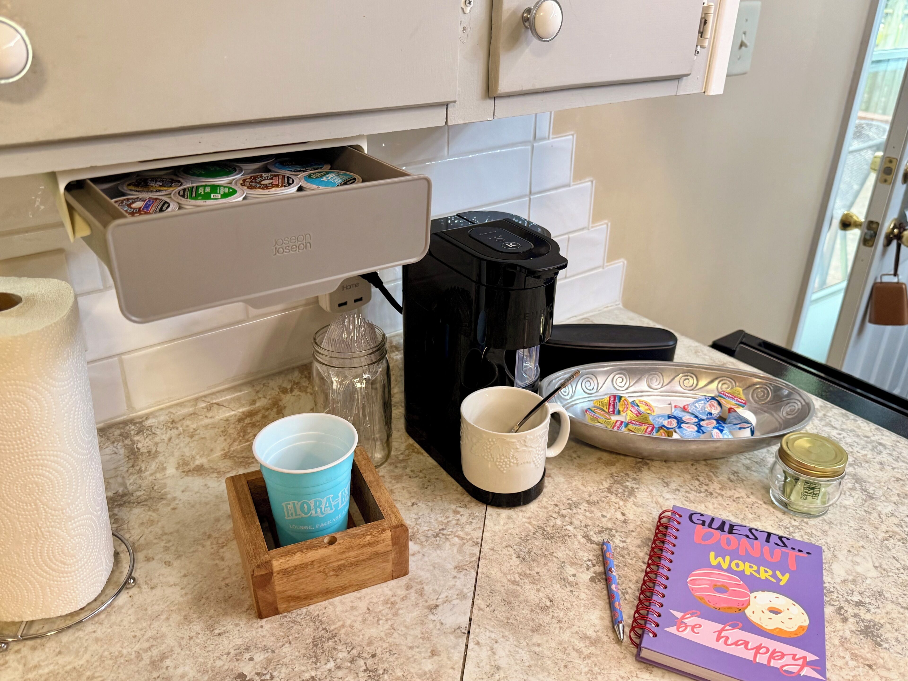 A kitchen counter with a coffee maker, a mug, a plate with snacks, a notebook, and a roll of paper towels.