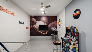 A hallway with a basketball and baseball sign, a ceiling fan, a TV screen, and a Pac-Man arcade machine. - Storey Lake Resort 545 (Orlando)