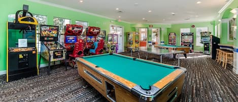 Une table de billard entourée de jeux d’arcade, dans une pièce aux murs verts et au tapis à motifs.
