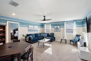Ein Wohnzimmer mit Ecksofa, einem Esstisch mit Stühlen, einem an der Wand montierten Fernseher und einem Deckenventilator.