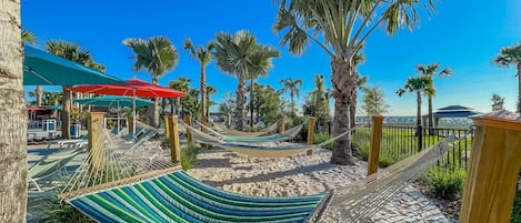 Una hamaca montada en una playa arenosa, con palmeras y sombrillas al fondo.