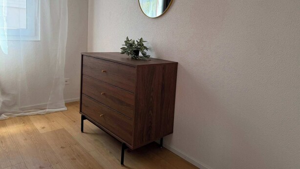 A room with a wooden dresser, a round mirror on the wall, and a window with sheer curtains.