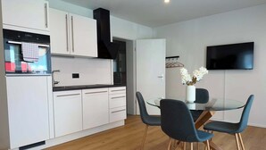 A modern kitchen with white cabinets, a black range hood, and a built-in oven. There is a dining area with a glass table and two chairs. A flat-screen TV is mounted on the wall.