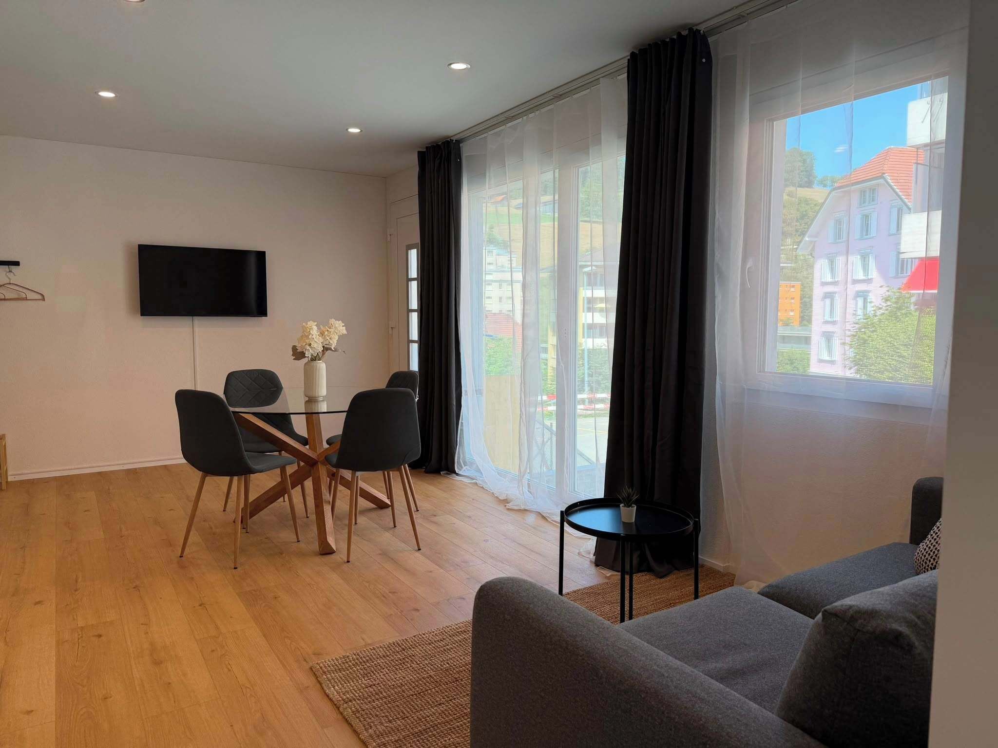 Une chambre moderne équipée d’une table à manger, de chaises, d’un téléviseur et d’une grande fenêtre avec des rideaux.