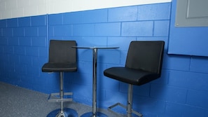 Dos sillas negras con patas metálicas y una pequeña mesa redonda entre ellas, frente a una pared azul con azulejos blancos.