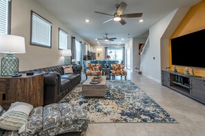 A spacious living room with a leather sofa, patterned ottoman, and a coffee table. There is a dining area with blue chairs and a kitchen in the background. - LNA3811 22034 - Five Bedroom Villa, Sleeps 12 (Orlando)