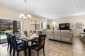 Un comedor con una mesa dispuesta para cuatro personas, un sofá, un televisor y vista a la piscina.