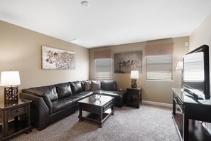 A living room with a leather sofa, a coffee table, and a TV stand. There are two lamps on side tables, and artwork is displayed on the walls. - Championsgate 994 - Six Bedroom Villa, Sleeps 14 (Orlando)