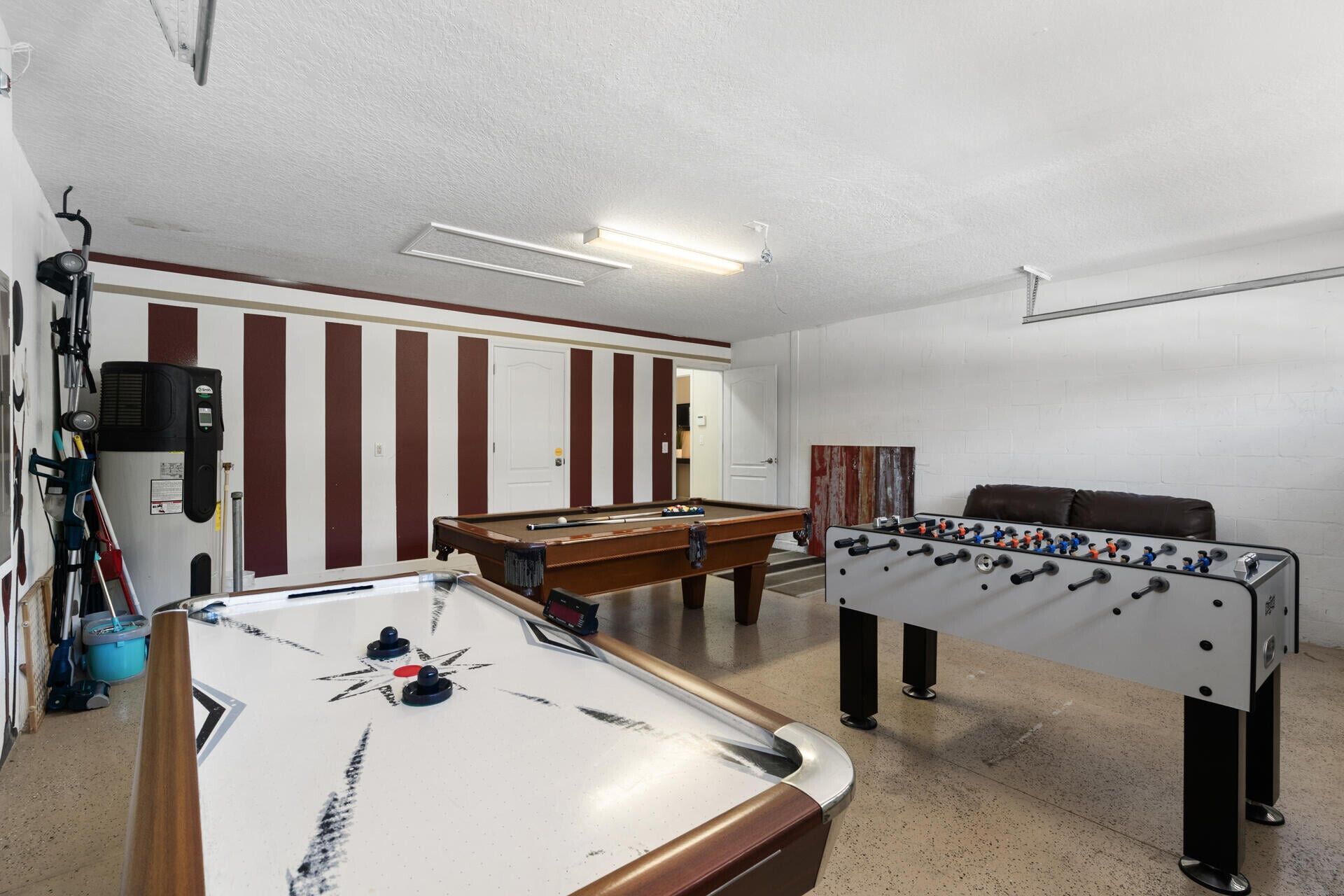 Une table de billard, une table de baby-foot et une table de jeu de fléchettes se trouvent dans une pièce aux murs rayés et dotée d’une porte.