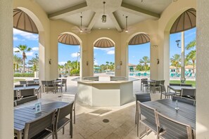 Ein Terrassenbereich im Freien mit Tischen und Stühlen, eine zentrale Bar sowie Blick auf einen Pool und Palmen.