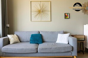 Ein graues Sofa mit drei Kissen, einem gerahmten abstrakten Kunstwerk, einem kleinen farbenfrohen Schild und einem Vintage-Radio auf einem Holzständer.