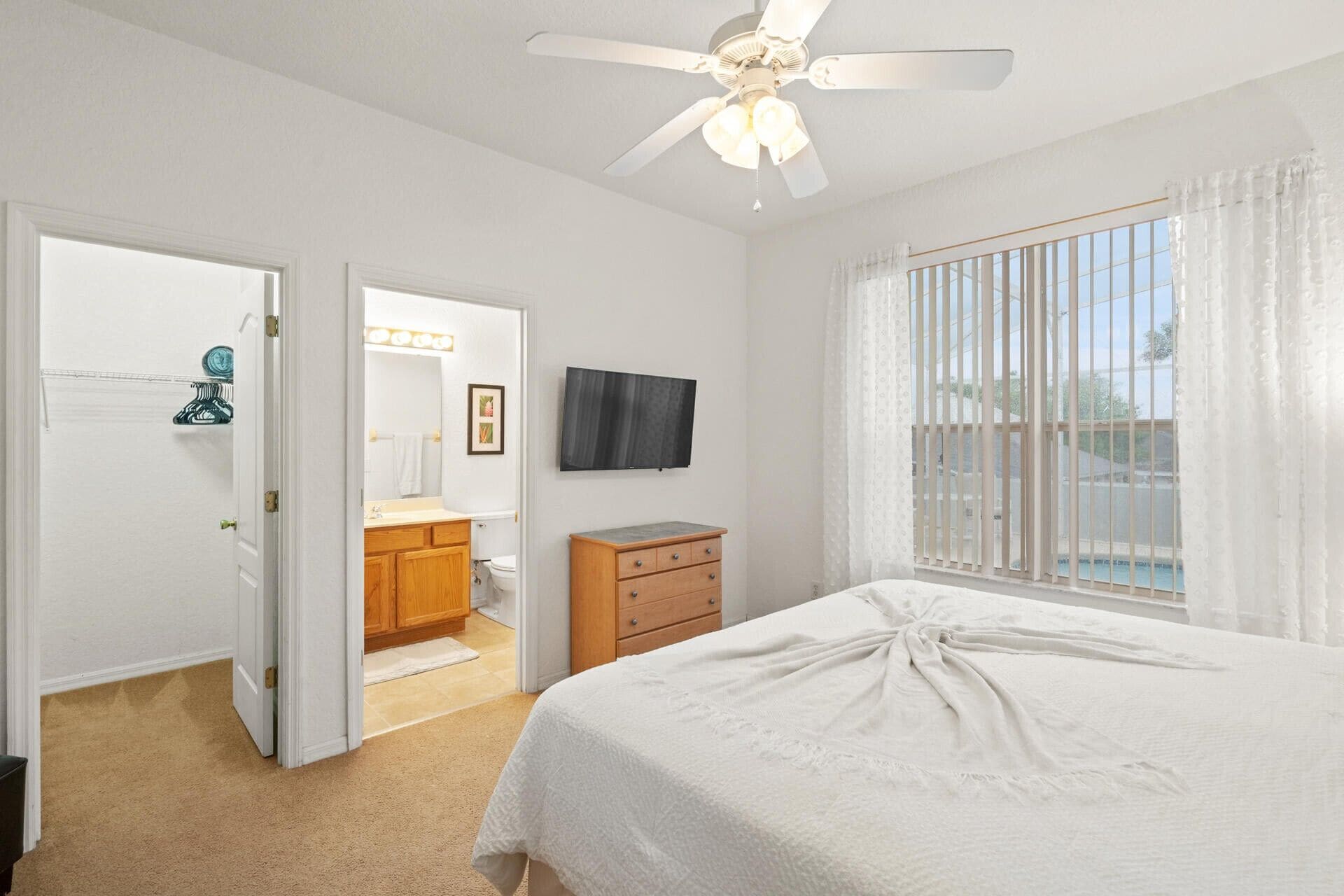 Une chambre à coucher avec un lit, un ventilateur de plafond, une télévision et une fenêtre avec des rideaux.