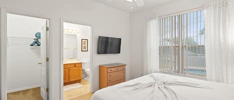 Une chambre à coucher avec un lit, un ventilateur de plafond, une télévision et une fenêtre avec des rideaux.