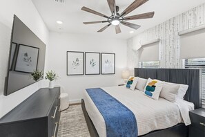 Ein Schlafzimmer mit einem Bett, Deckenventilator, Nachttisch, Lampe und gerahmten Bildern an der Wand.