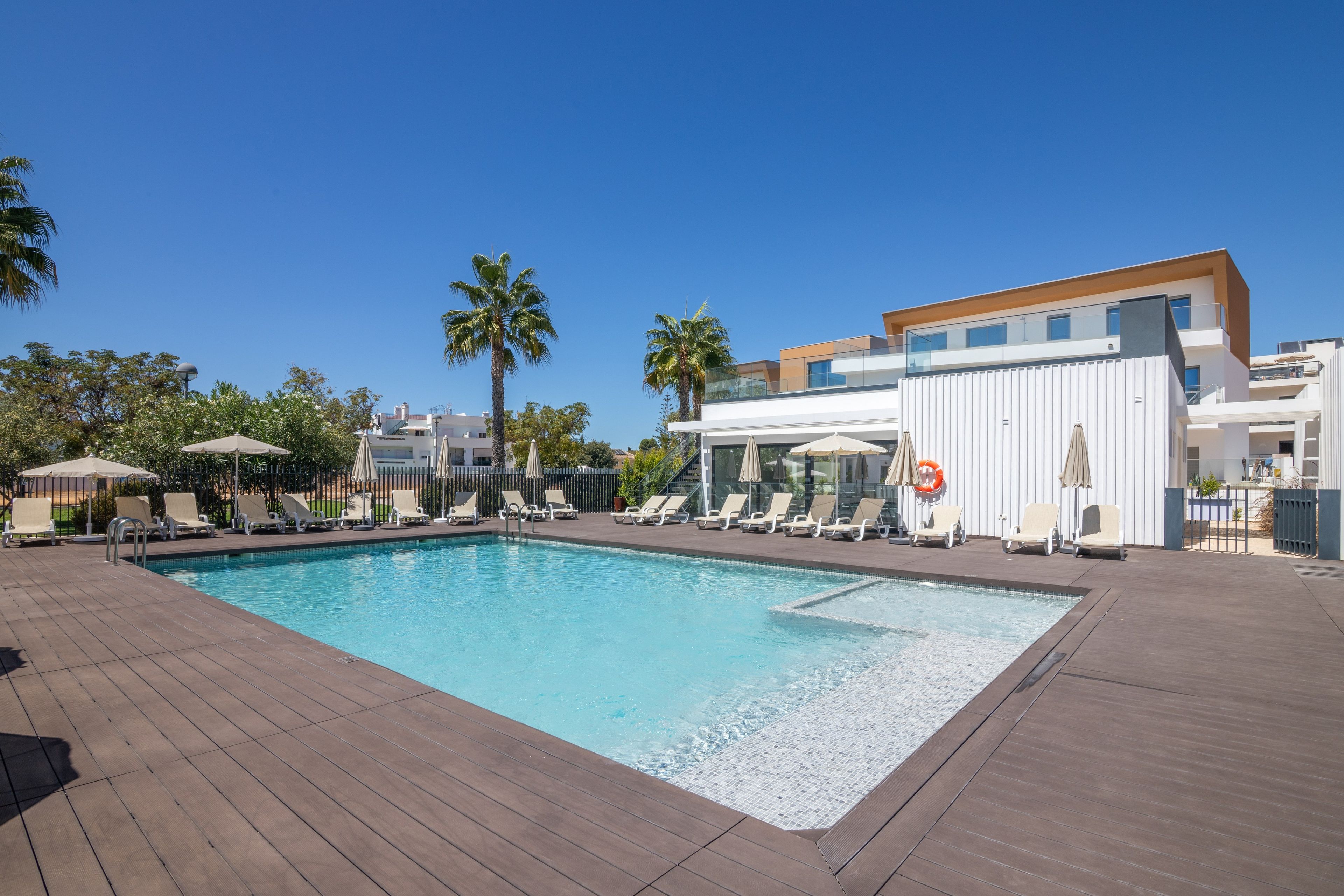 A swimming pool with lounge chairs and umbrellas, surrounded by palm trees and modern buildings.