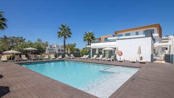 A swimming pool with lounge chairs and umbrellas, surrounded by palm trees and modern buildings.