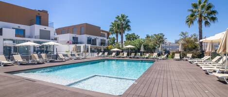 A pool area with lounge chairs and umbrellas, surrounded by buildings and palm trees.