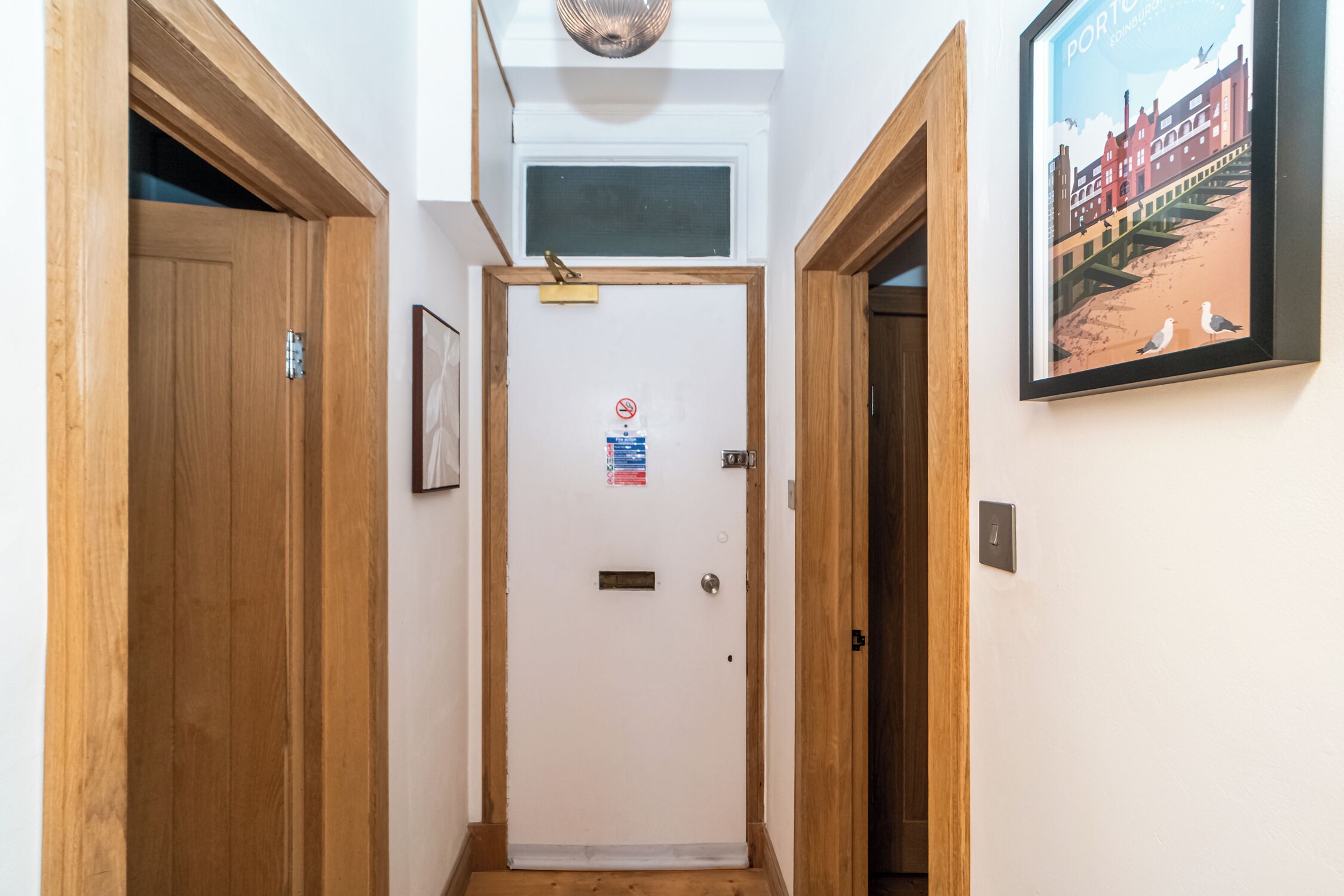 Un couloir étroit, avec des portes en bois de chaque côté, une porte blanche portant une inscription et un tableau encadré accroché au mur.