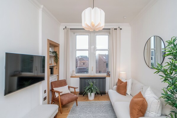 Un salon moderne avec un téléviseur à écran plat, un canapé, un fauteuil, une plante en pot et une grande fenêtre avec des rideaux.