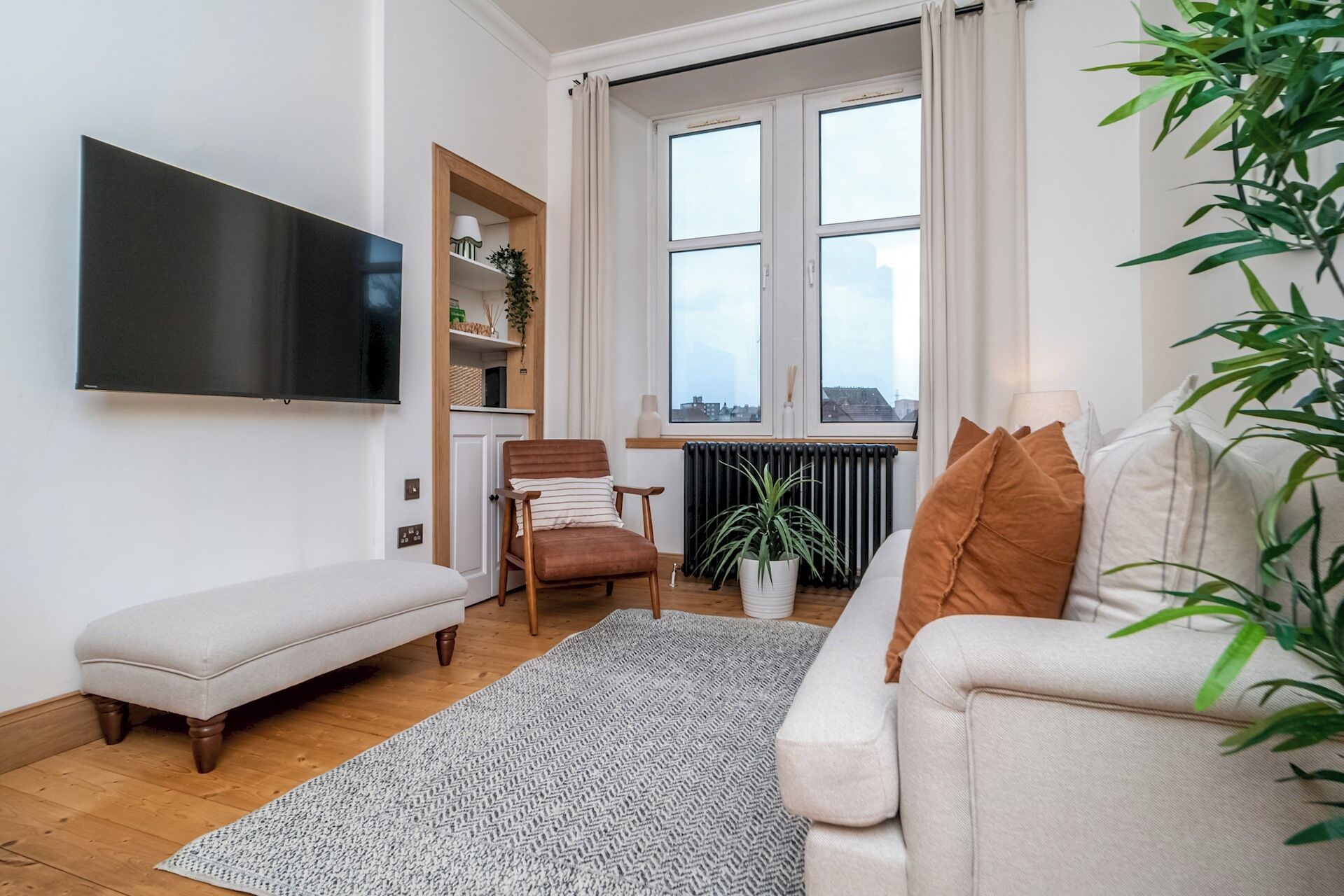 A living room with a sofa, a bench, a chair, a television, a radiator, and a window with a view.