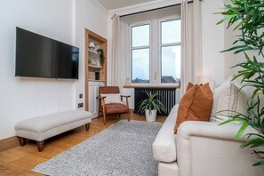 Een woonkamer met een bank, een lange bank, een stoel, een televisie, een radiator en een raam met uitzicht.