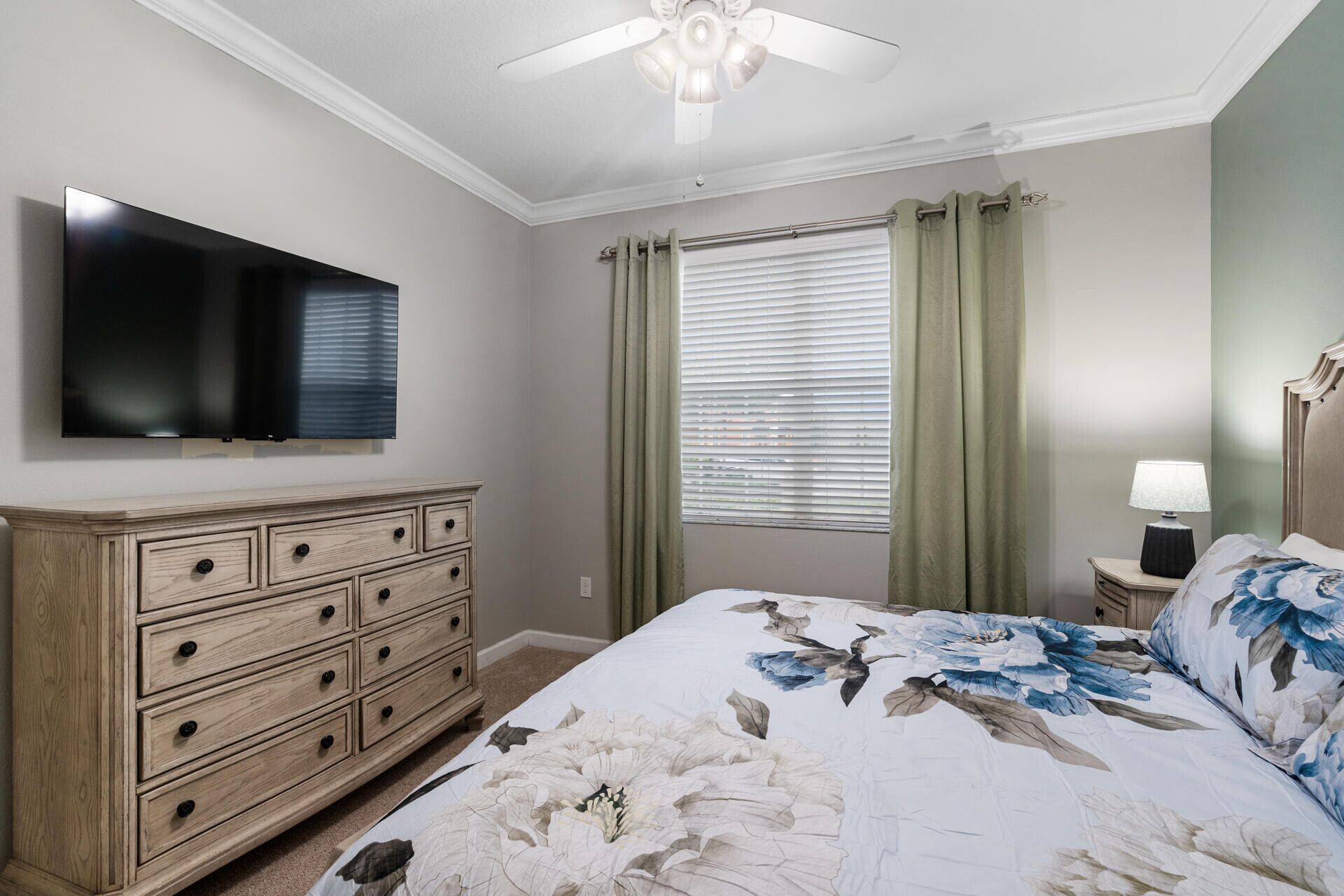 A bedroom with a bed, a dresser, a television, a window with curtains, and a ceiling fan.