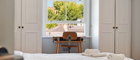 Ein Schlafzimmer mit Bett, Stuhl, Schreibtisch und einem Fenster mit Blick auf Grünflächen und Gebäude.