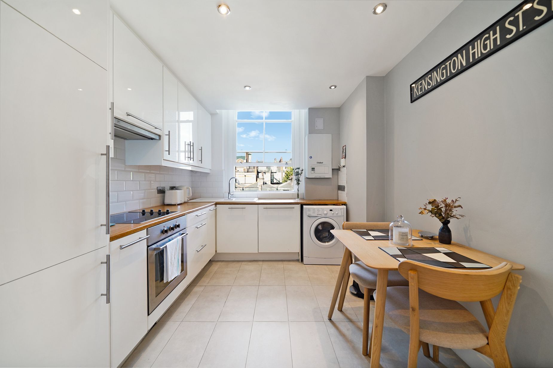 A modern kitchen with white cabinets, a wooden countertop, a built-in oven, a washing machine, and a dining area with a table and chairs.
