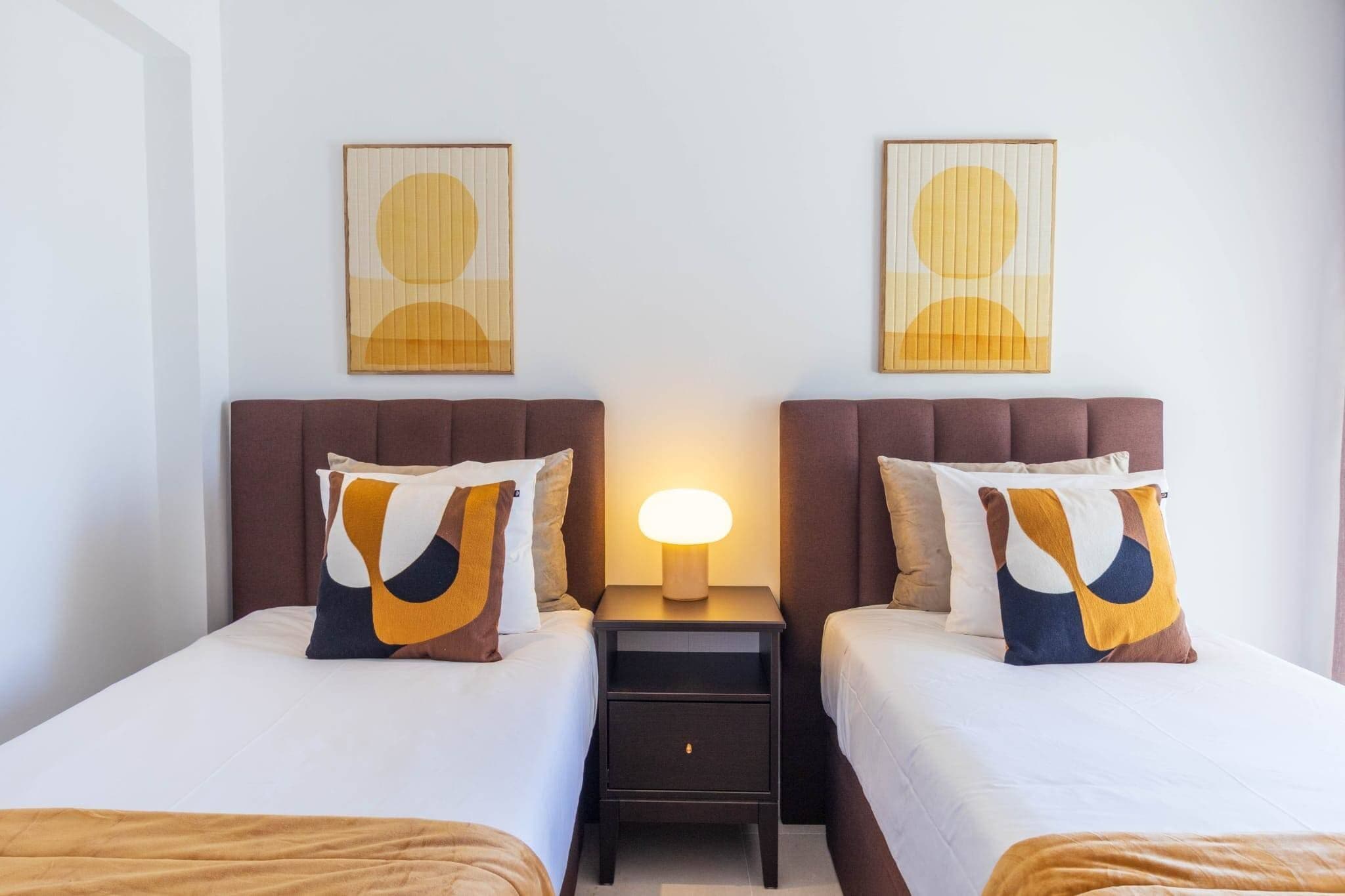 A hotel room with two beds, a nightstand with a lamp, and two wall-mounted artworks.