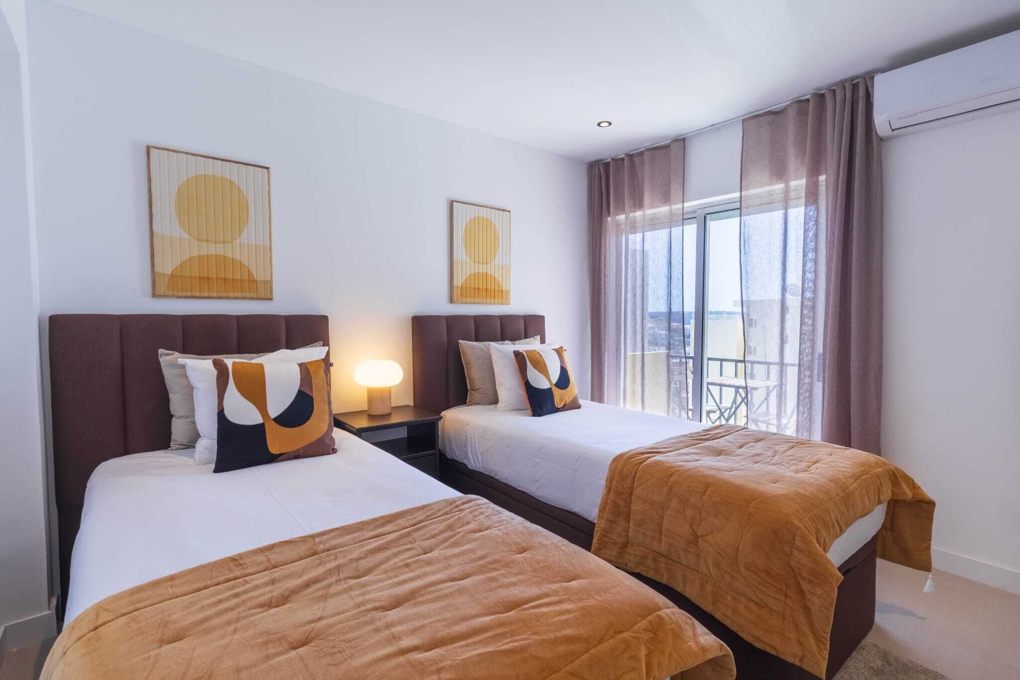 A hotel room with two beds, a nightstand with a lamp, and a balcony with a view.