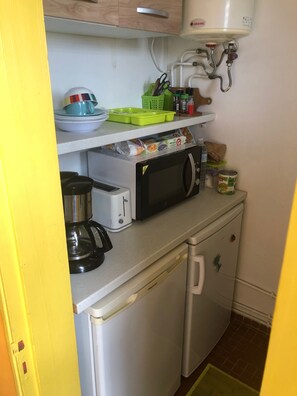 Et kompakt tekøkken med mikroovn, brødrister og kaffemaskine på bordet, et lille køleskab nedenunder, og en hylde med tallerkener og en grøn kurv.