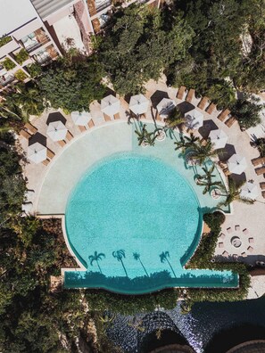 Ein Luftbild eines Resorts mit einem großen, runden Pool, umgeben von Liegestühlen und Sonnenschirmen, mit Palmen und einem Gebäude im Hintergrund.