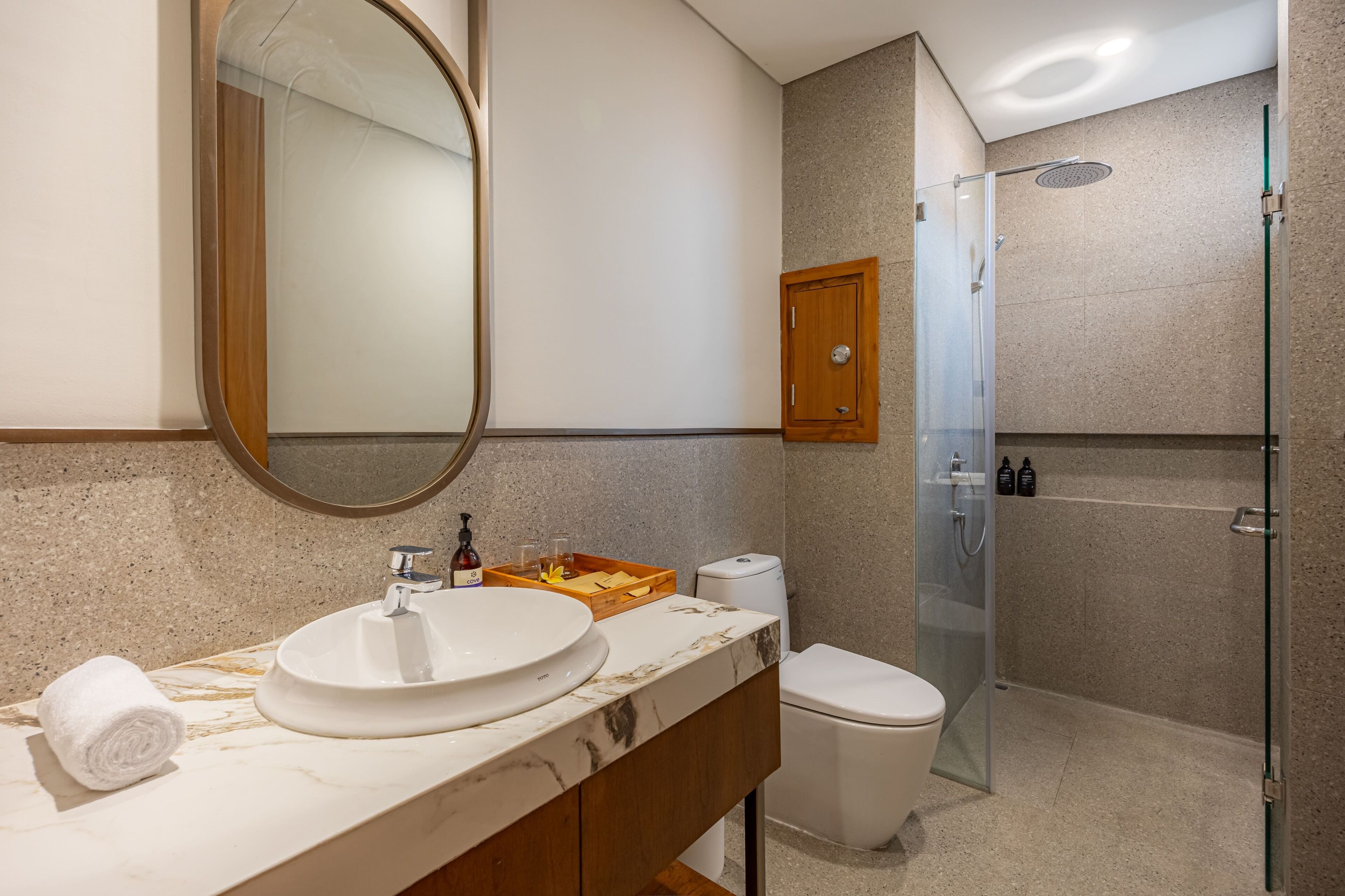 Un baño con un lavamanos ovalado grande, un inodoro, una ducha con mampara de vidrio y un armario de madera.