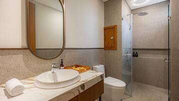 Un baño con un lavamanos ovalado grande, un inodoro, una ducha con mampara de vidrio y un armario de madera.